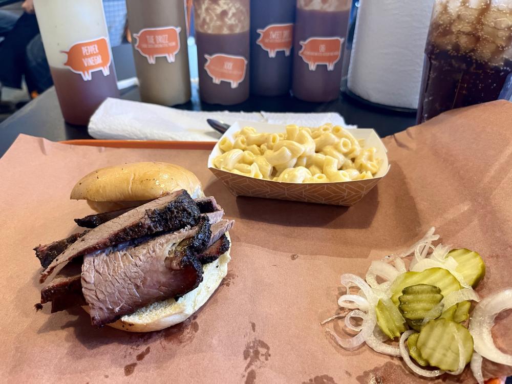 Barbecue brisket sandwich with mac and cheese, pickles, and onions on brown paper.