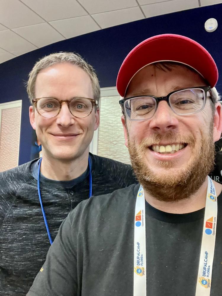 Two people smiling indoors, wearing glasses and casual clothes.