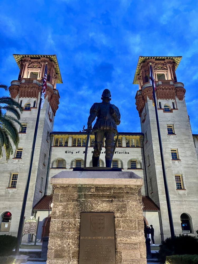 Statue of a soldier in front of a historic building at dusk, with palm trees and flags.