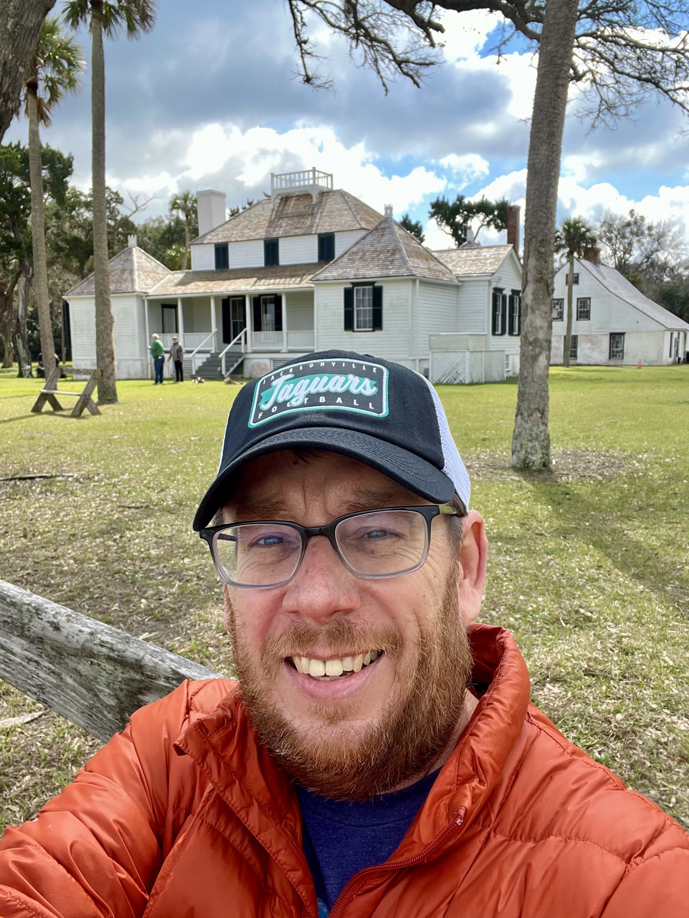 Man in glasses and cap smiling in front of a historic white house on a sunny day.