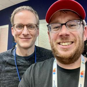 Two people smiling indoors, wearing glasses and casual clothes.