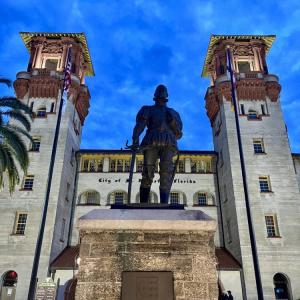 Statue of a soldier in front of a historic building at dusk, with palm trees and flags.