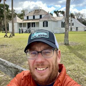 Man in glasses and cap smiling in front of a historic white house on a sunny day.